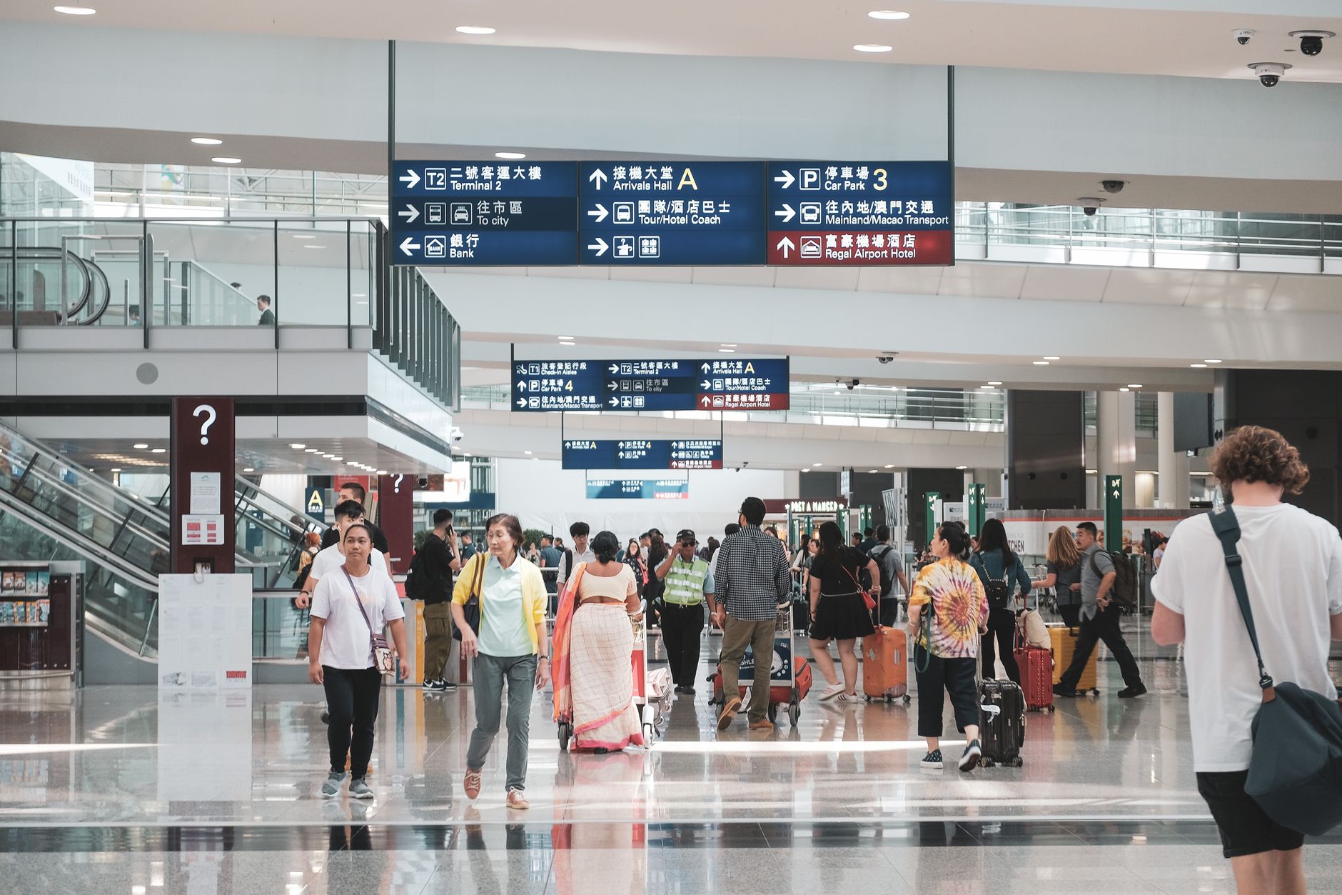 Airport terminal interior with directional signs and crowds of passengers with luggage moving through concourse.