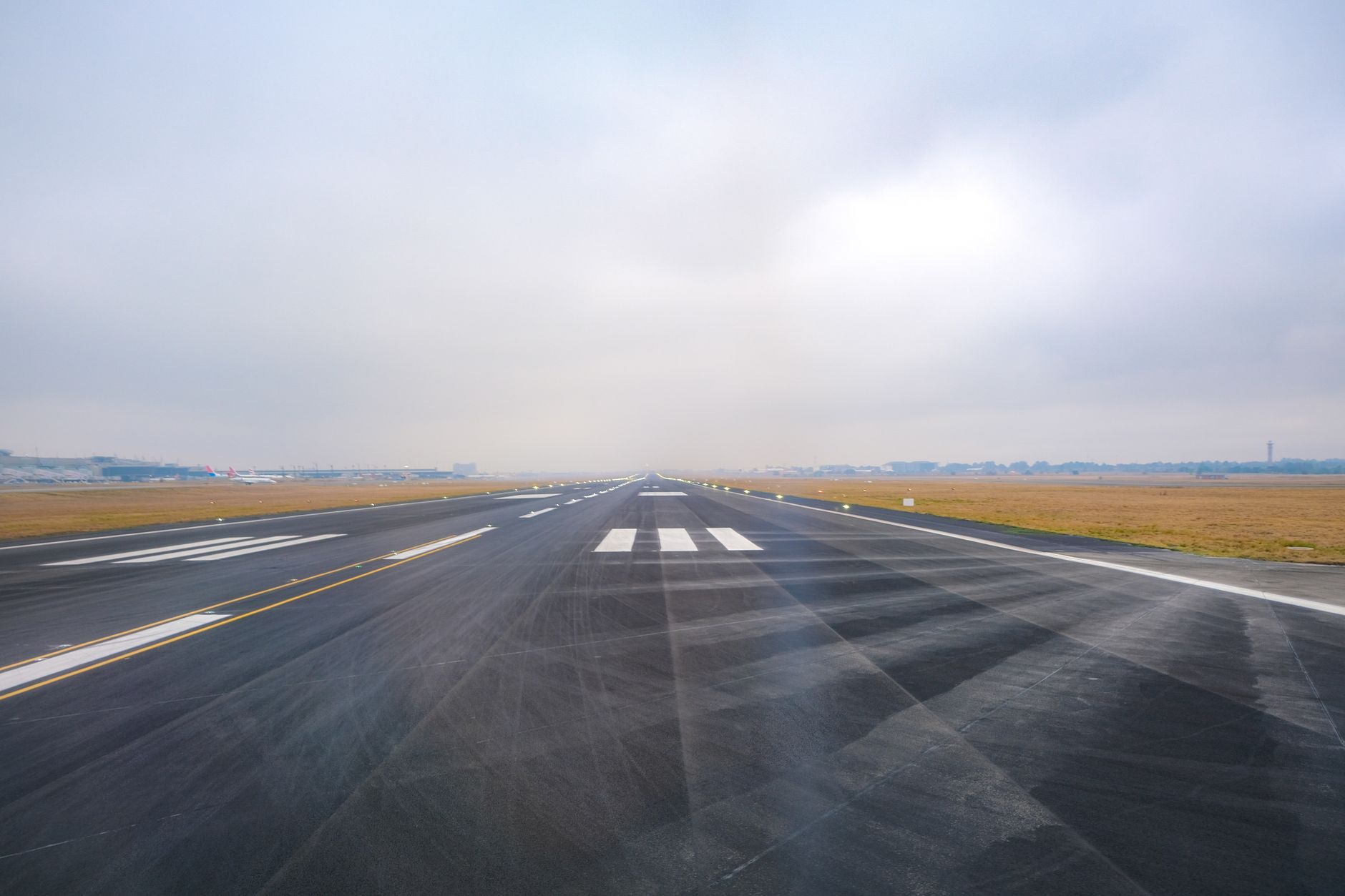 Empty airport runway with white centerline markings and approach threshold stripes extending into distance under overcast sky