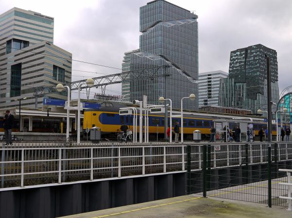 Yellow and blue passenger train at a modern station platform with city buildings behind, representing Birmingham-Manchester Airport rail service