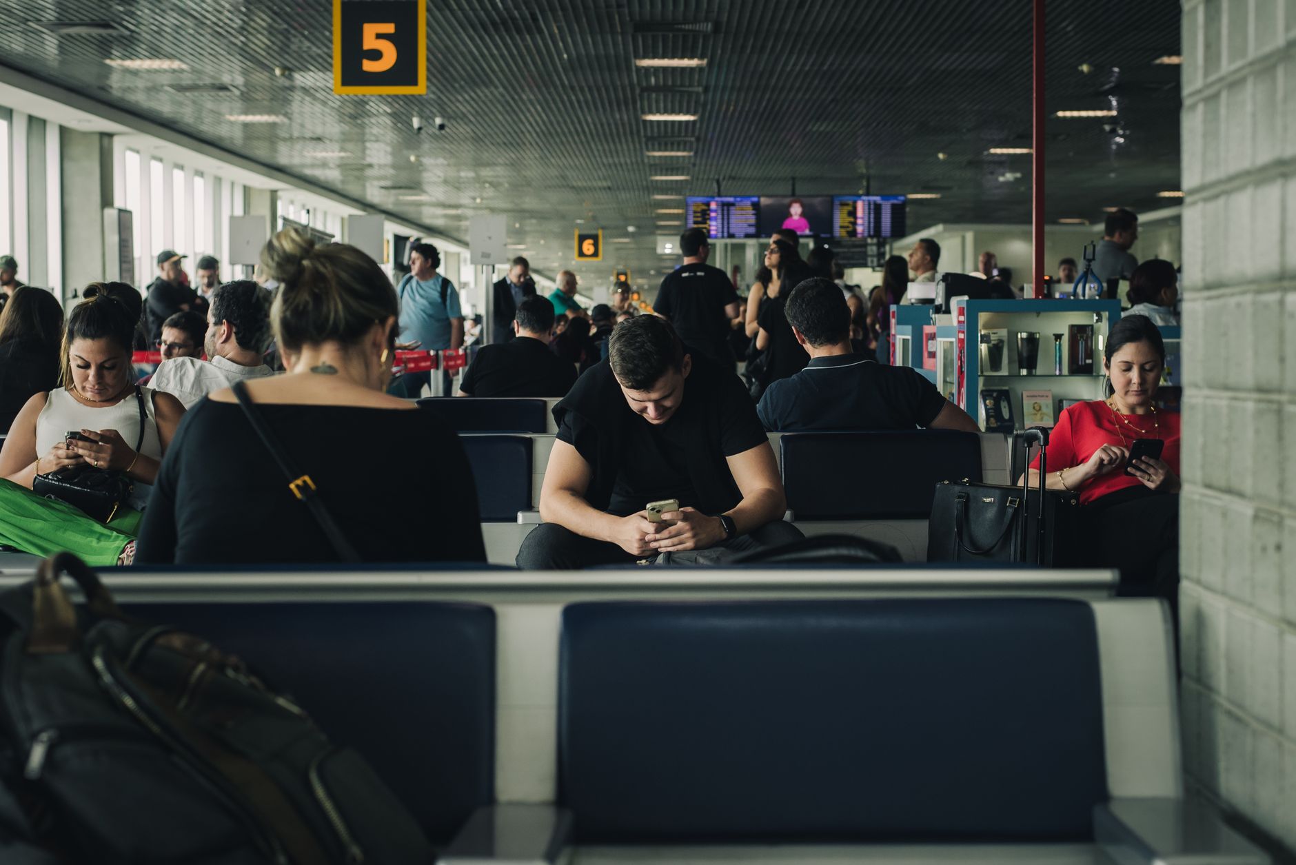 Passengers waiting in airport terminal with gate numbers 5 and 6 visible, departure boards overhead